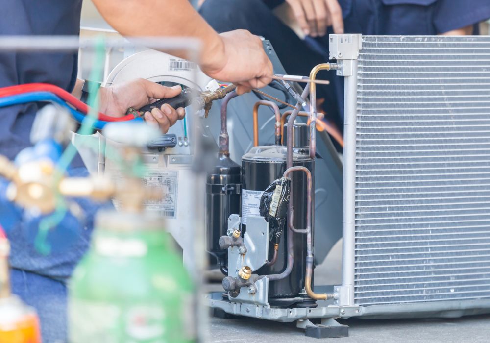 Close-up of a technician repairing an air conditioning unit, manipulating copper tubing and tools near the compressor and condenser.