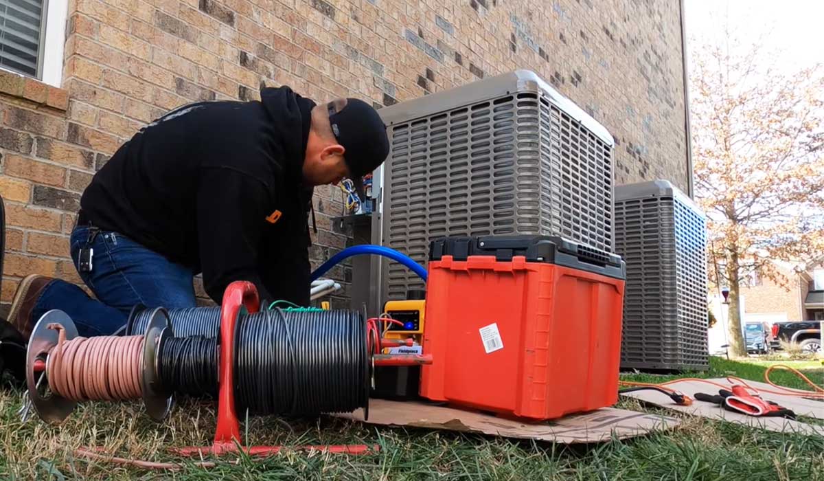 A technician kneels beside outdoor HVAC units with tools, hoses, and cables, representing hands-on service and real-world factors affecting HVAC repair cost.